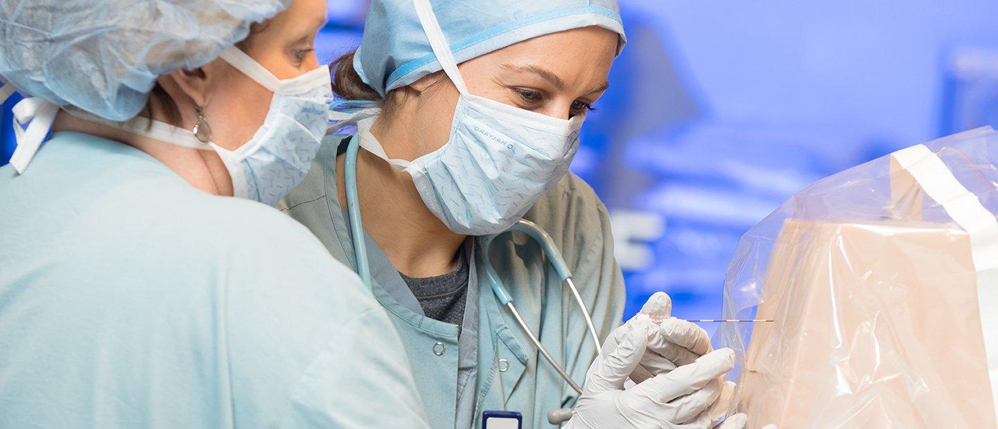 two women in surgical gowns and masks poking an object in a plastic cover